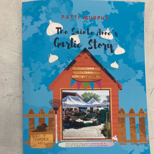 La couverture bleue d'un livre de TS intitulé « Livre - The Sainte Anne's Garlic Story » de Patti Murphy comporte une illustration d'un hangar en bois avec une fenêtre représentant une scène de marché. Sous le titre, on peut lire, en texte anglais, « From Farm to Table Festival ». La couverture comprend également des illustrations d'ail et des graphiques de terre de jardin.