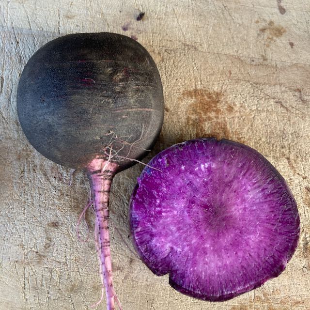 Un Radis d&#39;hiver Améthyste Noir - semences bio de Tourne-Sol est posé à côté d&#39;une coupe transversale de lui-même, révélant un intérieur violet vibrant. Ces légumes reposent sur une surface en bois texturée.