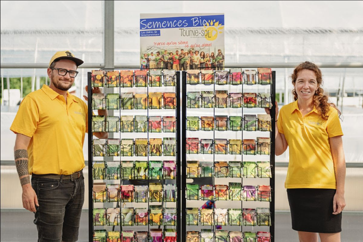 Deux personnes en chemise jaune se tiennent à côté d&#39;une exposition colorée de Don de Semences. Le panneau indique « Tourne-Sol » et le décor intérieur a une atmosphère de serre, soulignant l&#39;aspect organique.