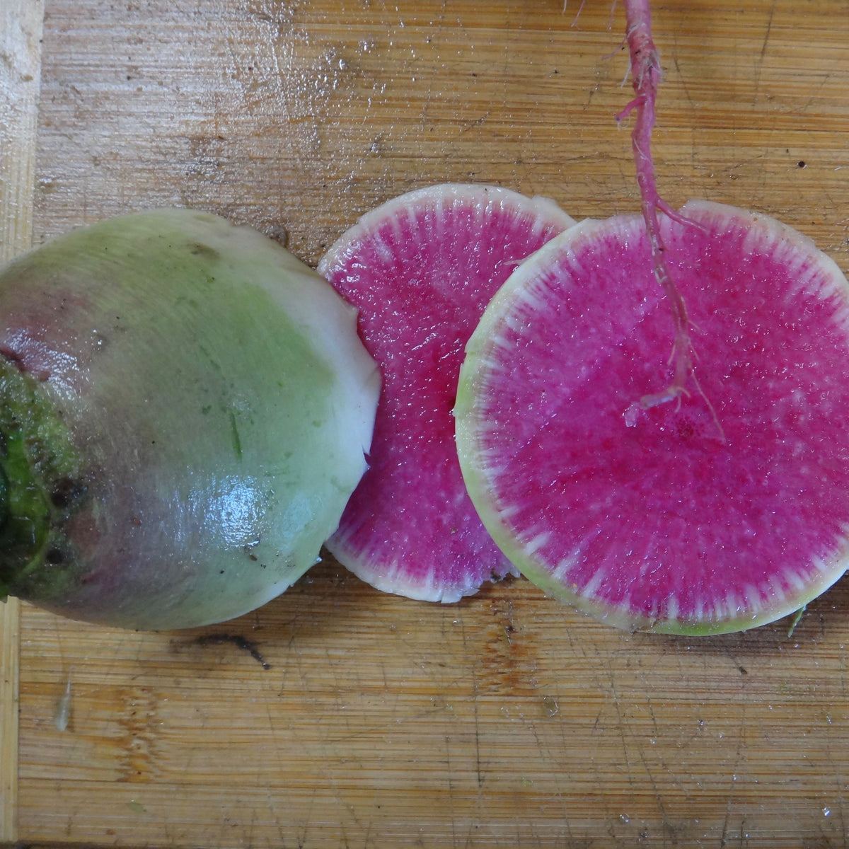 Un Radis Melon d&#39;Eau - semences bio de Tourne-Sol entier et deux morceaux tranchés reposent sur une planche à découper en bois, présentant un design rond avec une peau blanche à l&#39;extérieur et une chair rose vif à l&#39;intérieur.