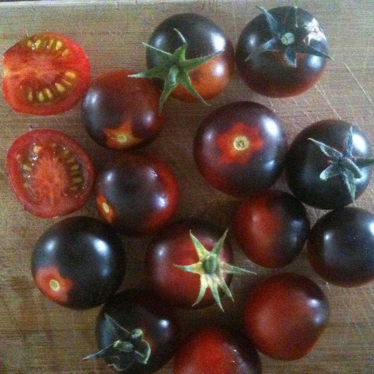 Une collection de tomates bleues cerises Dancing with Smurfs de Tourne-Sol, aux tons violet foncé et rouge, est posée sur une surface en bois. Leur peau bleu/mauve brille et deux tomates sont coupées en deux pour montrer leur intérieur juteux.